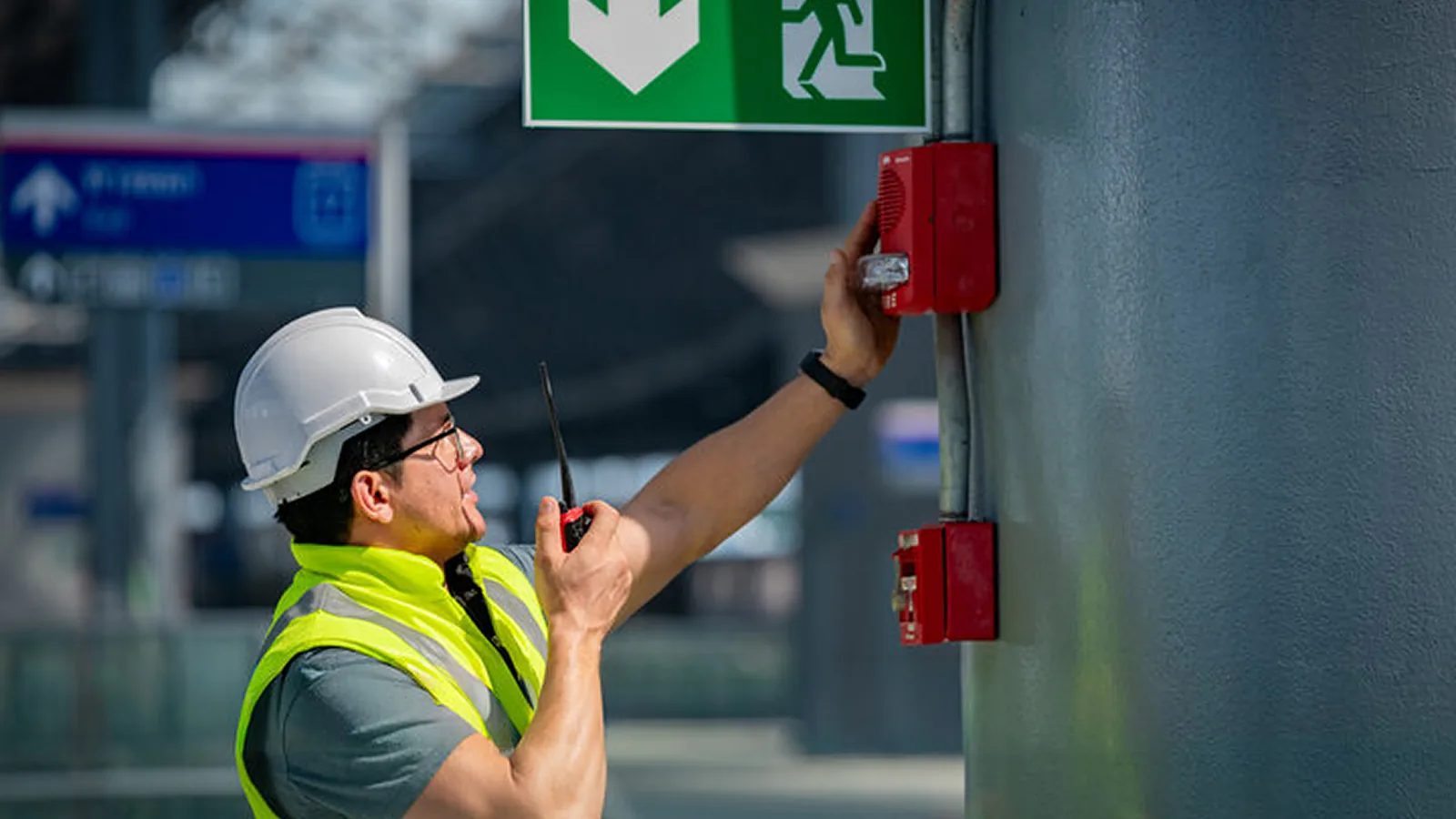Technician in hard hat and safety vest testing a wall-mounted fire alarm